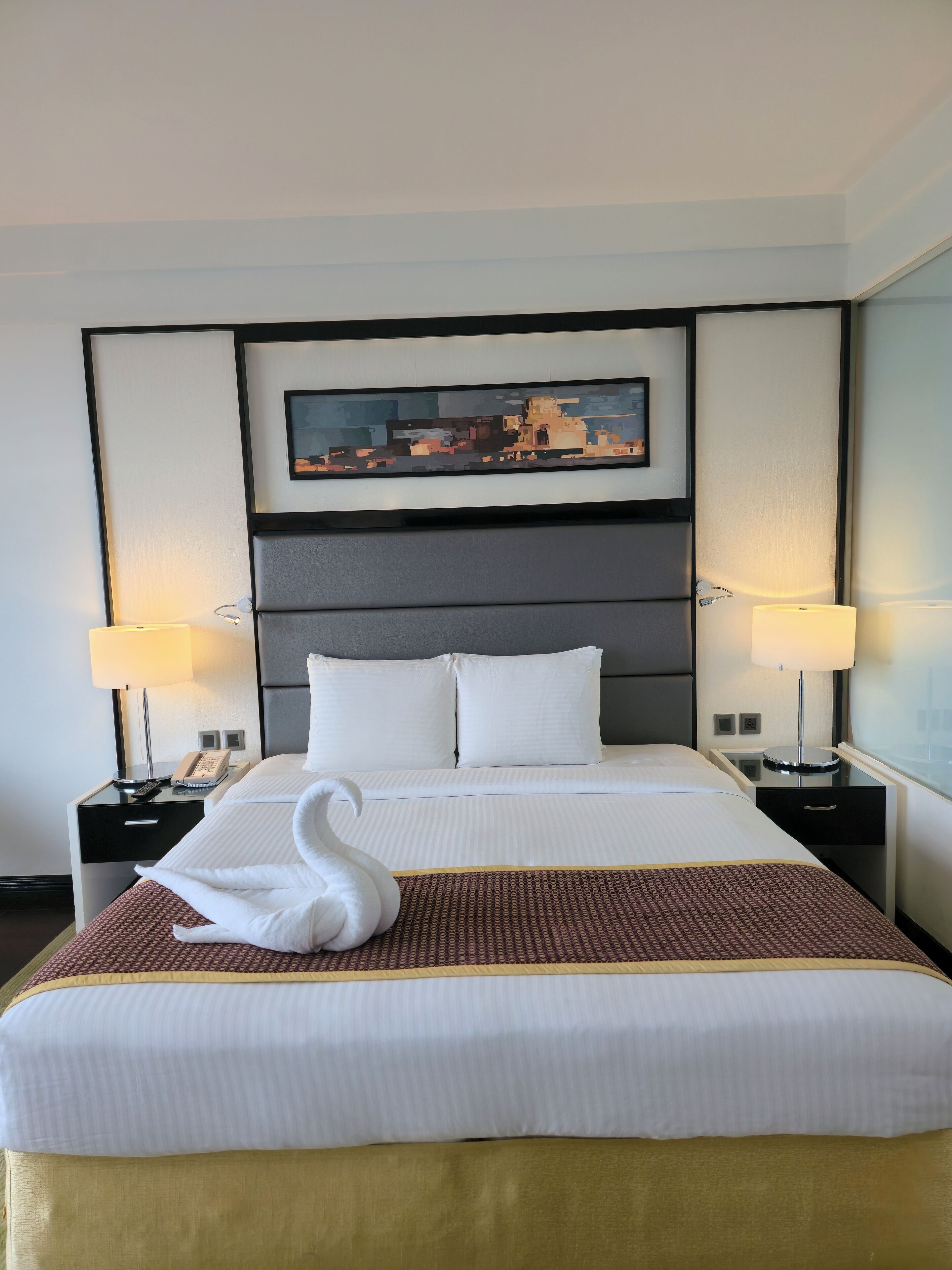 A hotel room with a large bed, two bedside tables, a wall-mounted painting, and a swan-shaped pillow.