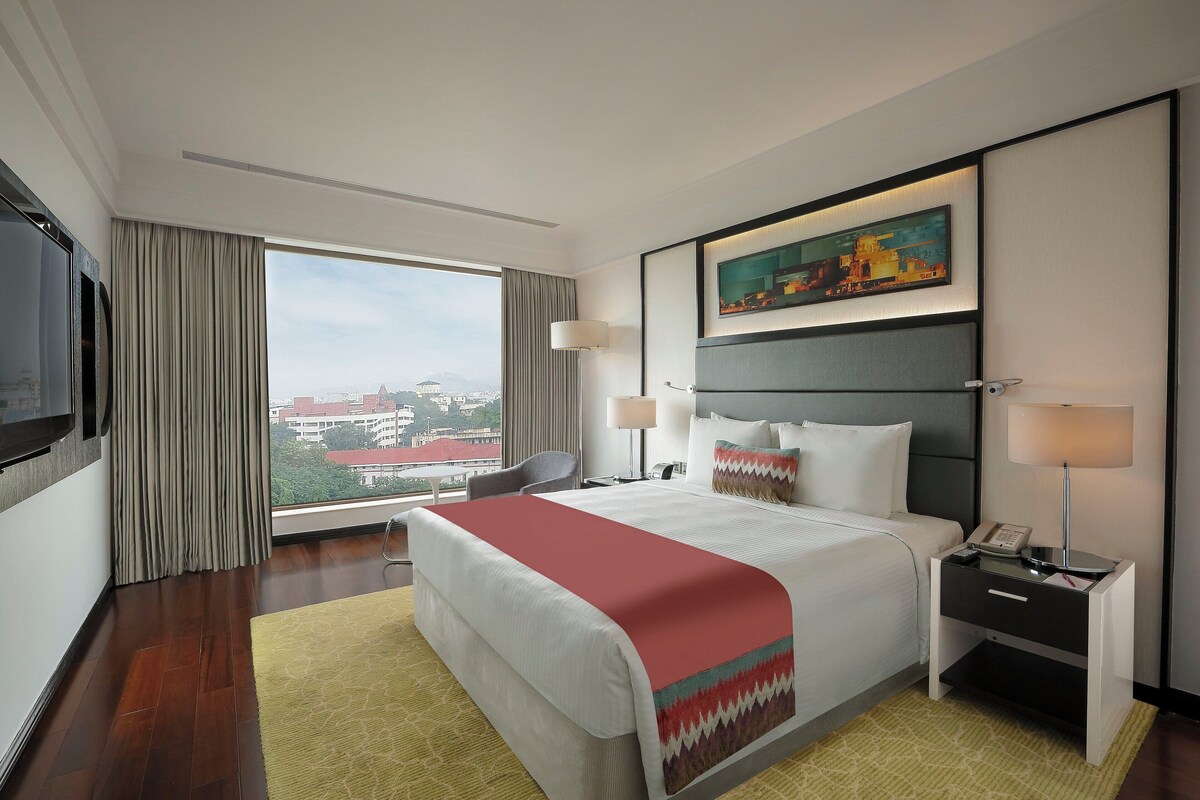 Une chambre d’hôtel avec un grand lit, un bureau avec un téléphone, une chaise, une télévision et une fenêtre donnant sur la ville.