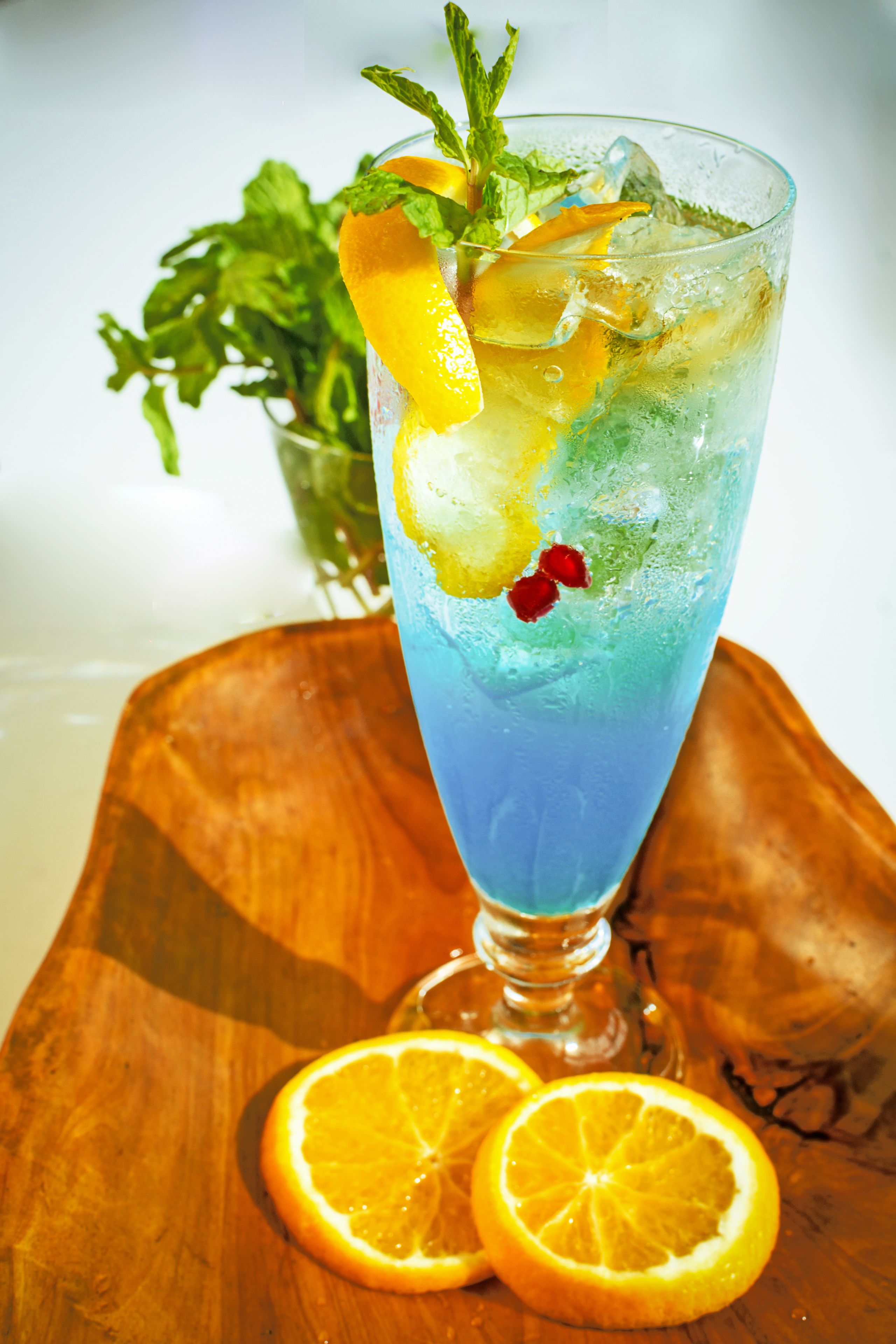 Un vaso alto con una bebida azul, decorado con rodajas de naranja y hojas de menta, sobre una tabla de madera.