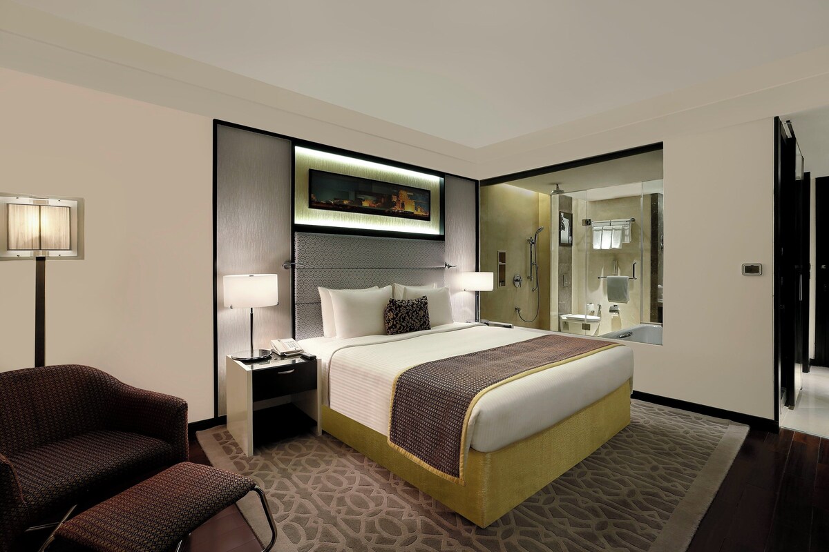 Une chambre d’hôtel avec un lit, des lampes de chevet, une chaise, un bureau avec un téléphone, un tableau au-dessus du lit et une salle de bain visible à travers une porte en verre.