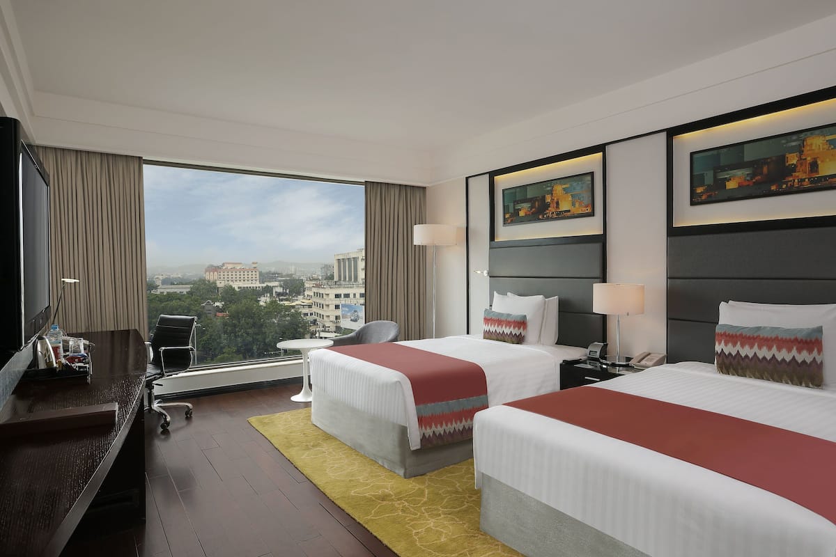 A hotel room with two beds, a desk, a chair, a small table, and a large window offering a city view.