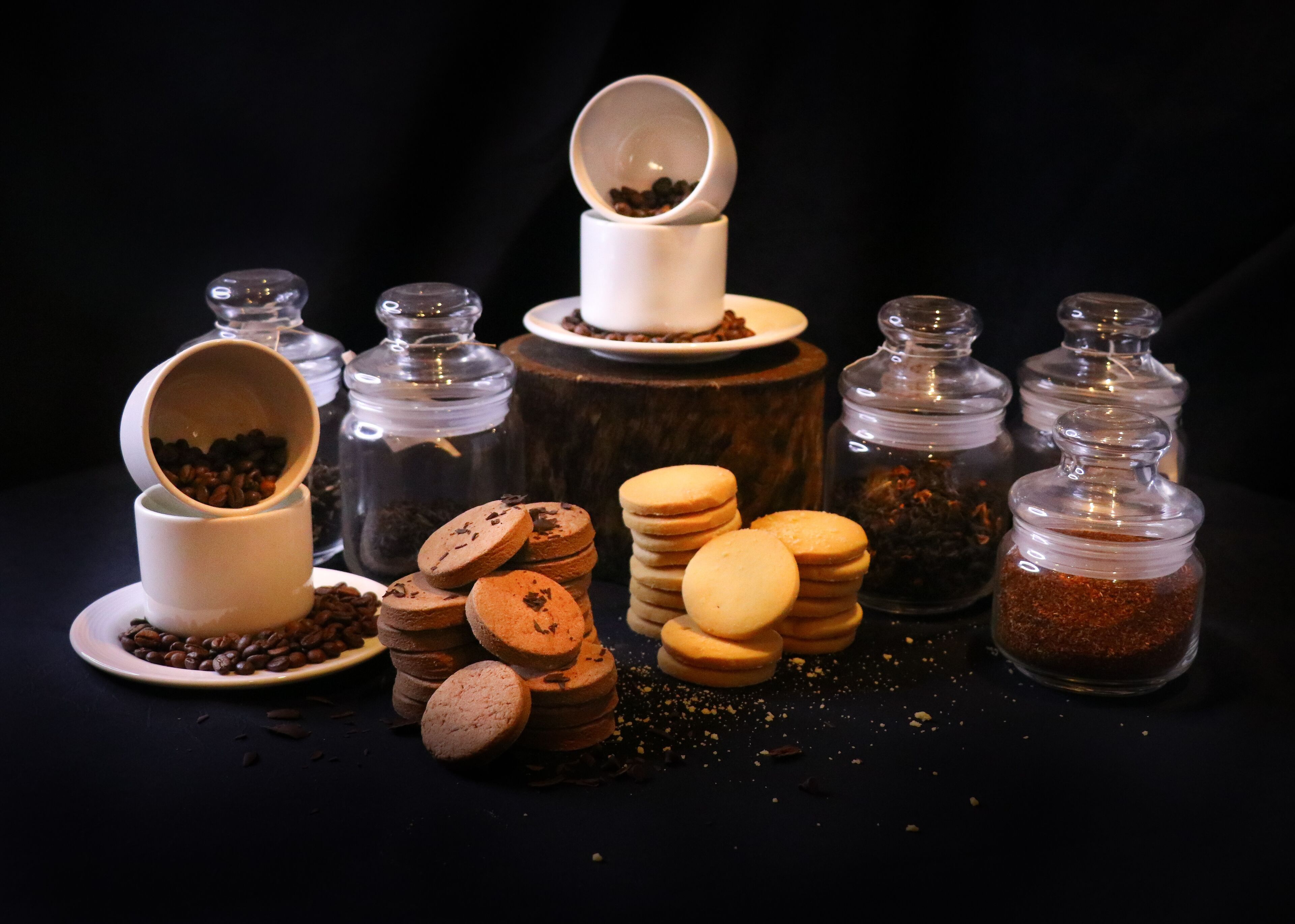 Verschiedene Lebensmittel und Behälter, darunter Kekse, Kaffeebohnen und Gewürze, auf einer dunklen Oberfläche angeordnet.