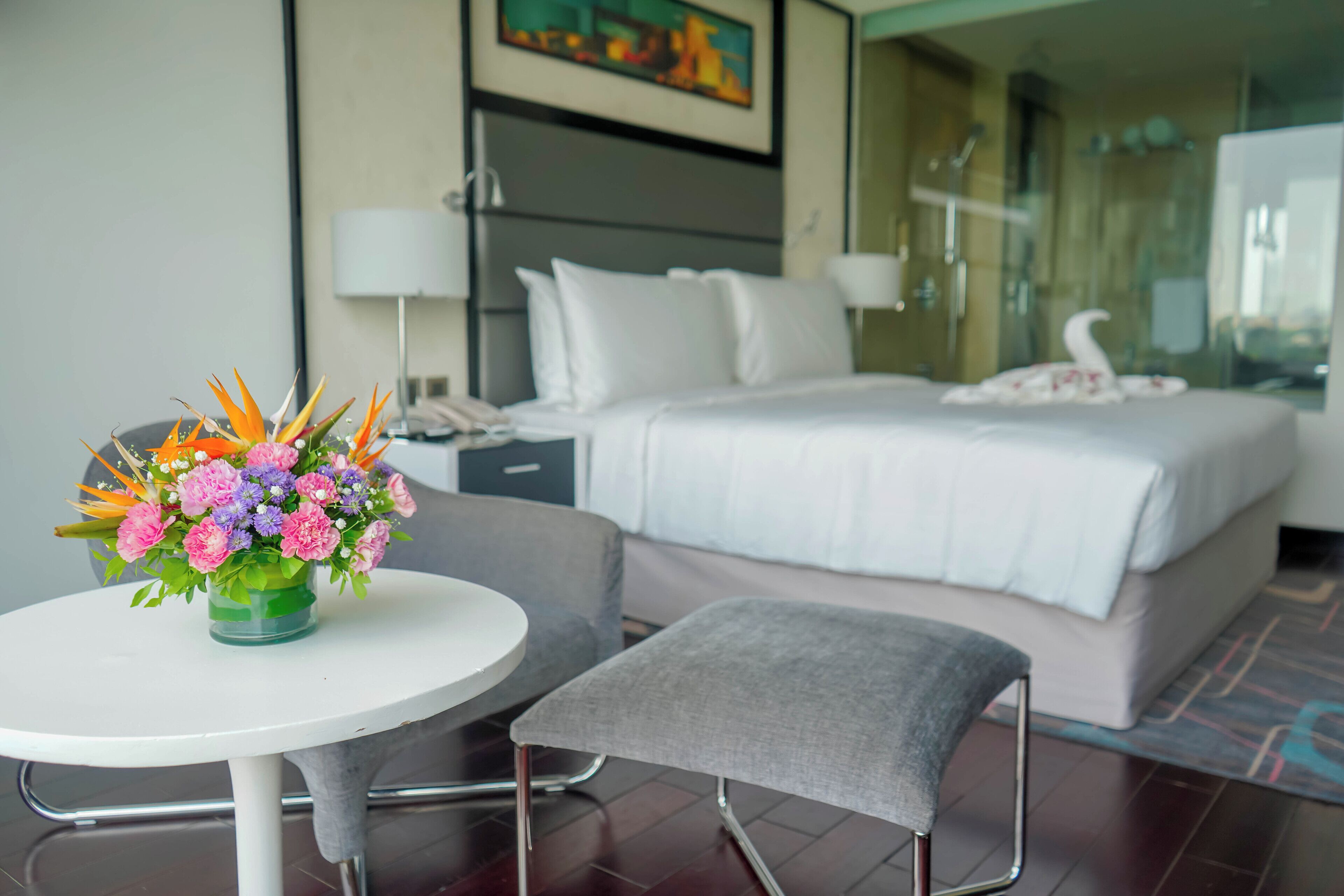 Una habitación de hotel con una cama, una mesita de noche con un jarrón de flores, una silla y un ventanal grande.
