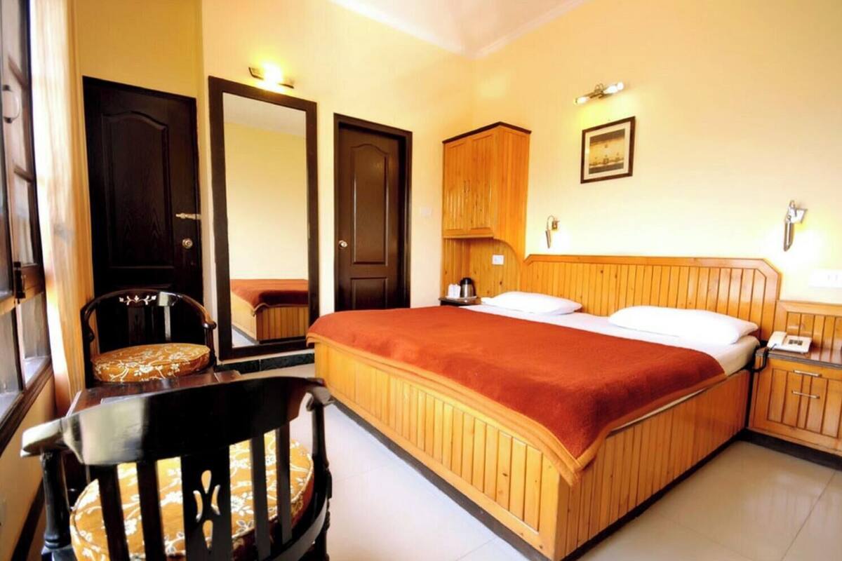 A bedroom with a wooden bed, a chair, a nightstand, a mirror, and a wall-mounted light fixture.