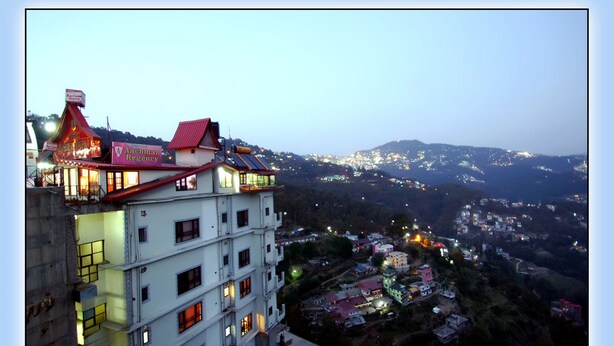 Un edificio de varios pisos con techo rojo y ventanas iluminadas, con vistas a un valle con casas y luces dispersas.