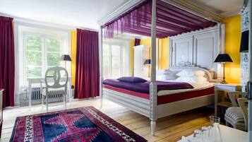A four-poster bed with a canopy, a large window with curtains, a chair, and a table with glasses and a pitcher.