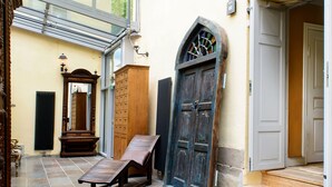 Un pasillo estrecho con techo de vidrio, bancos de madera, una puerta grande en arco y una escalera que sube a un nivel superior.