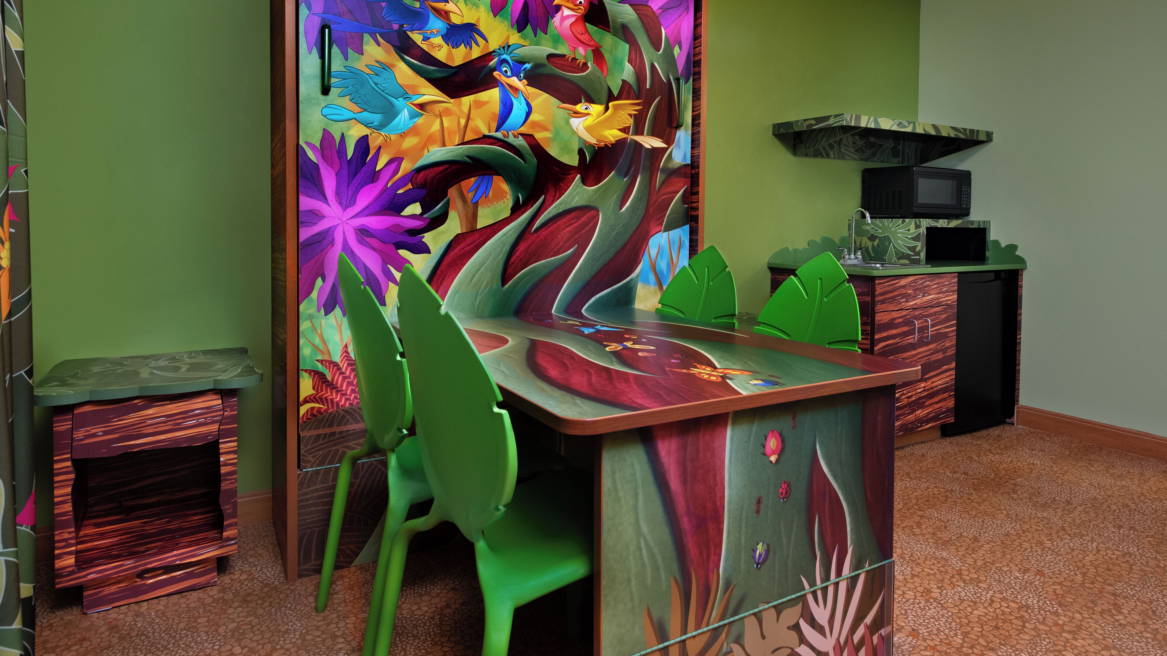 A room with a wooden desk, green chairs, a colorful mural, a microwave, and a small cabinet.