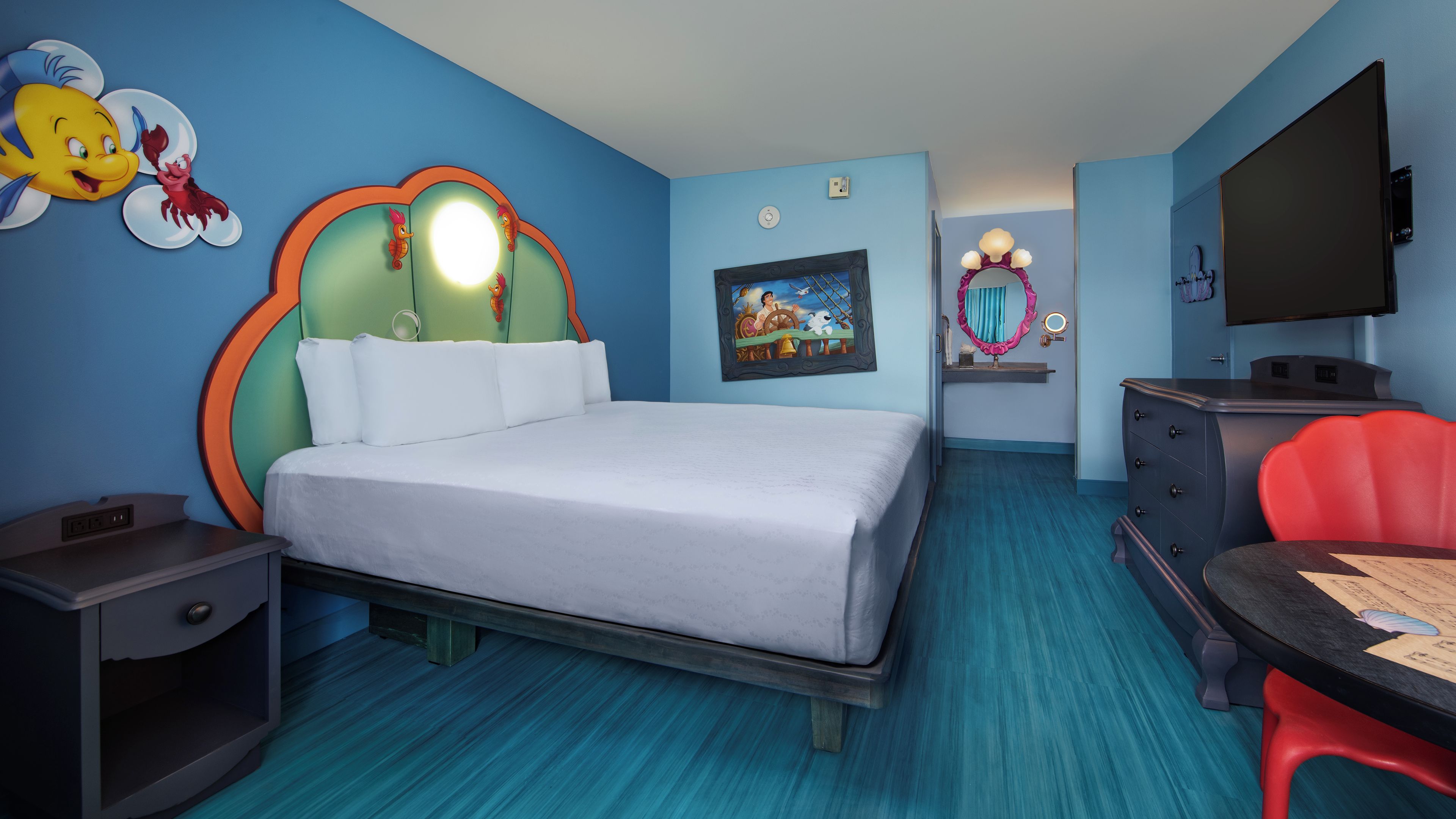Una habitación de hotel con cama, escritorio, televisor y una pared decorada con personajes de caricatura.