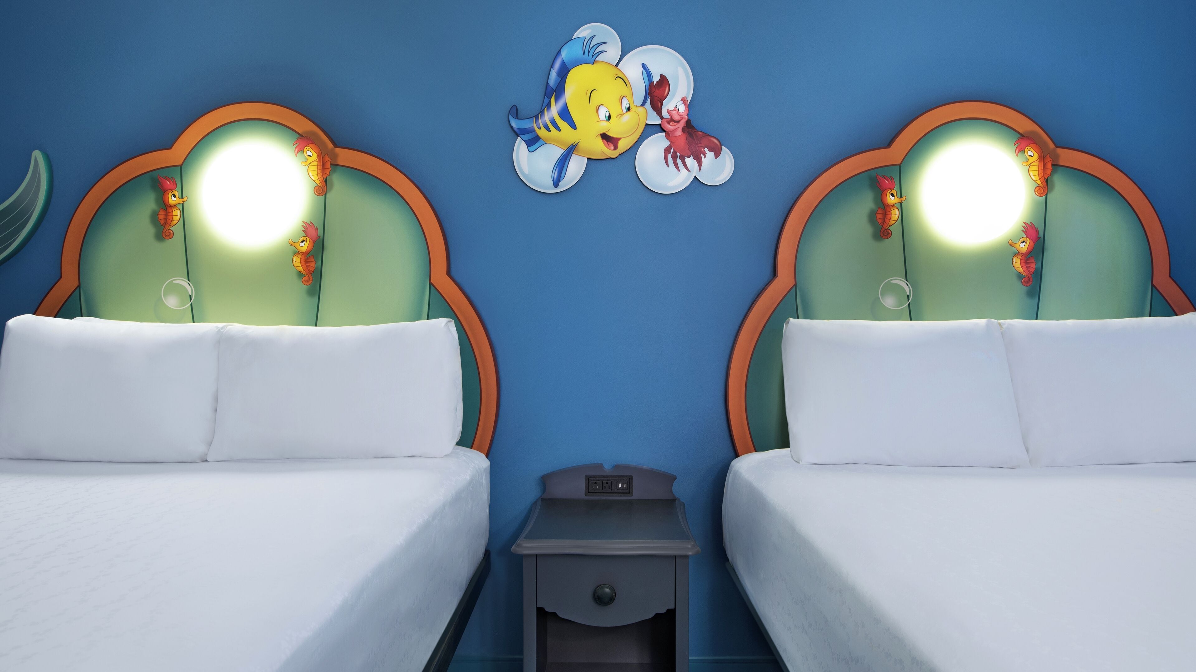 Ein Hotelzimmer mit zwei Betten, jedes mit einem Kopfende, das einen Cartoon-Charakter zeigt. Dazwischen befindet sich ein kleiner Nachttisch.