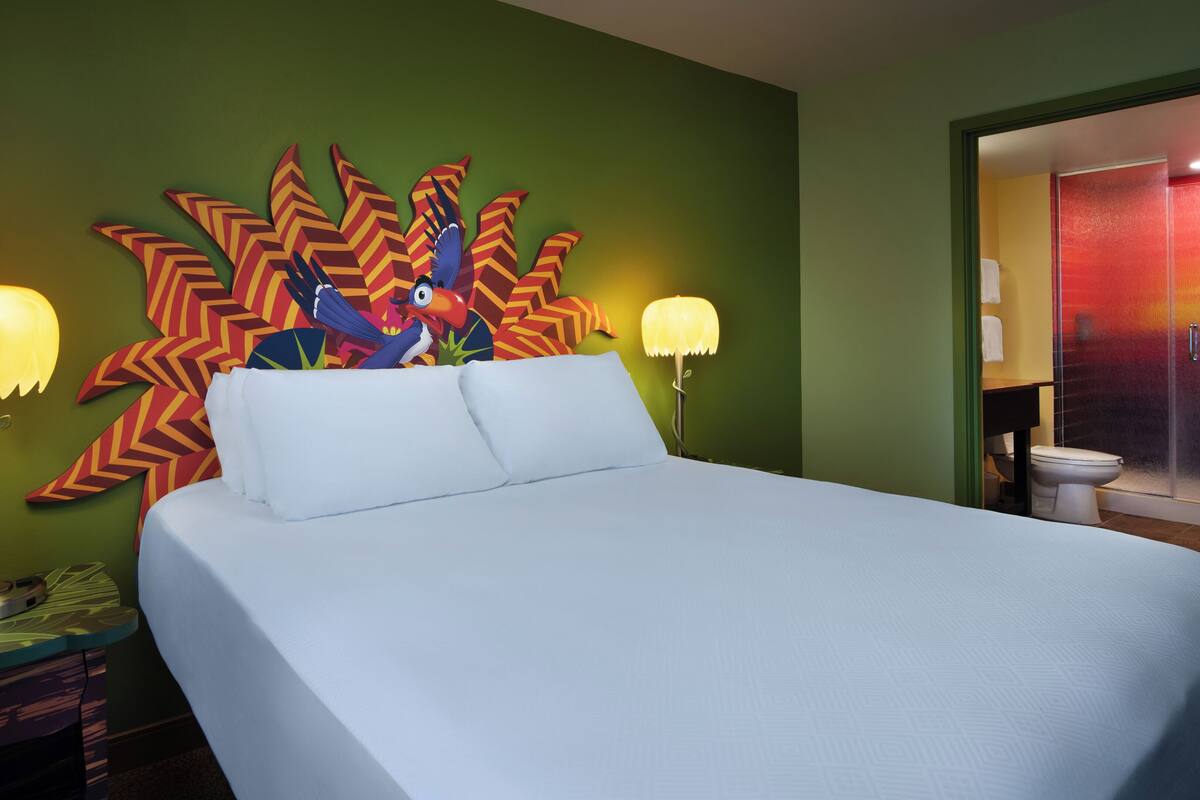 A hotel room with a bed, two bedside lamps, a colorful wall decoration, and a bathroom visible through an open door.