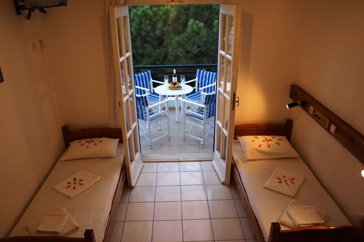 Quarto com duas camas, uma mesinha com cadeiras e vista para um jardim através de uma porta aberta.