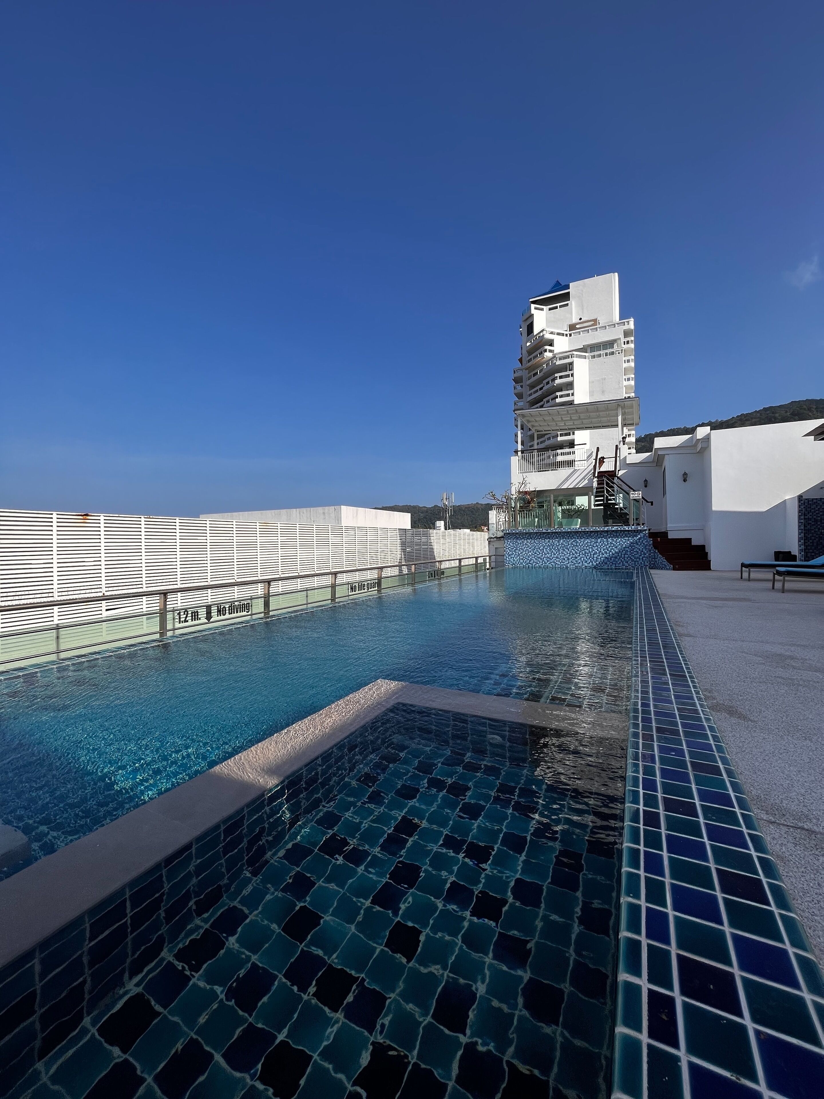 Ein Pool auf dem Dach mit blauem Himmel, ein weißes Gebäude mit Balkonen und ein Schild, das eine Tiefe von 12 Metern angibt.