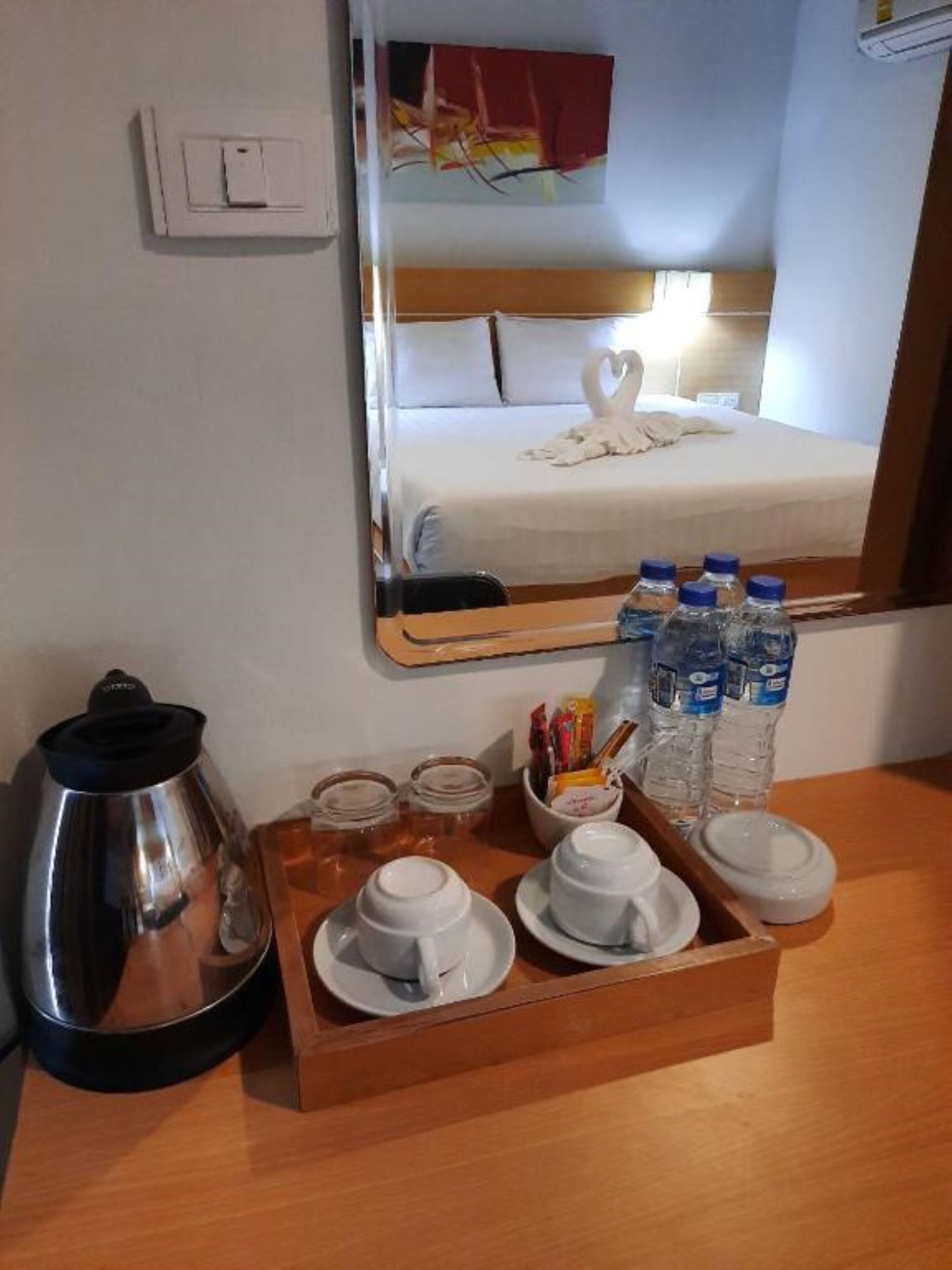 A hotel room with a bed, a mirror reflecting the bed, a tray with cups, a kettle, and bottled water.