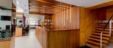 Un vestíbulo de hotel moderno con un mostrador de recepción de madera, una escalera y un área de comedor al fondo.