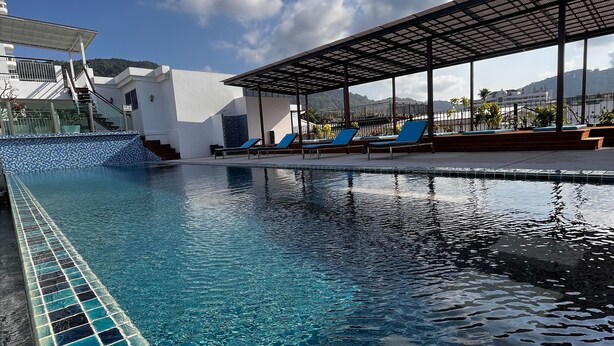 A swimming pool with clear blue water, a covered seating area with blue chairs, and a white building in the background. - Aspery Hotel (Patong)