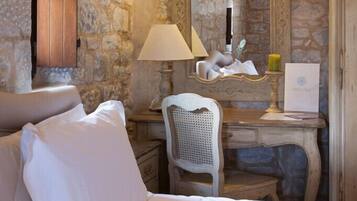 A bedroom with stone walls, a wooden ceiling, a bed with white linens, a desk with a lamp, and a mirror.