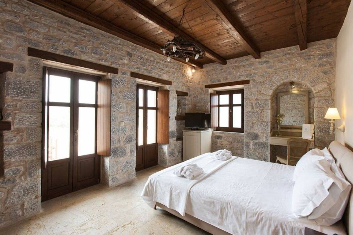 A bedroom with stone walls, wooden beams, a bed, a TV, and a desk with a chair.