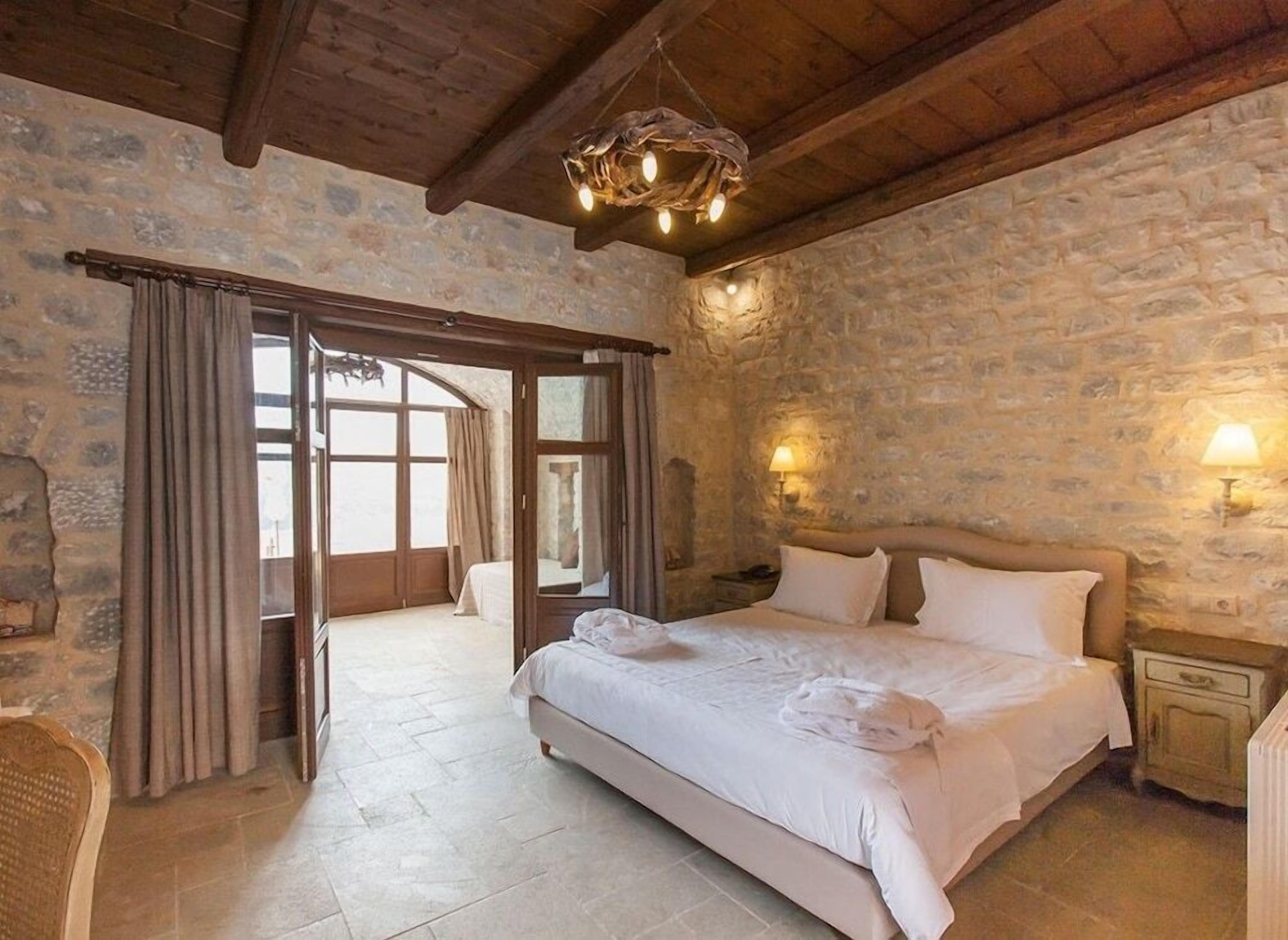 A bedroom with a stone wall, wooden ceiling beams, a large bed, and a wooden chair.