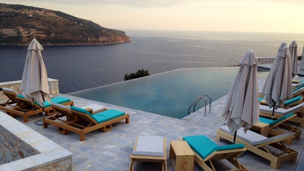 A poolside area with wooden loungers and umbrellas, overlooking a body of water and a hilly landscape.