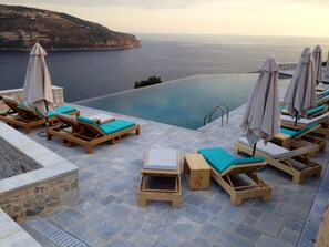 Un espace aménagé au bord de la piscine, avec des chaises longues en bois et des parasols, offrant une vue sur une étendue d’eau et un paysage vallonné.