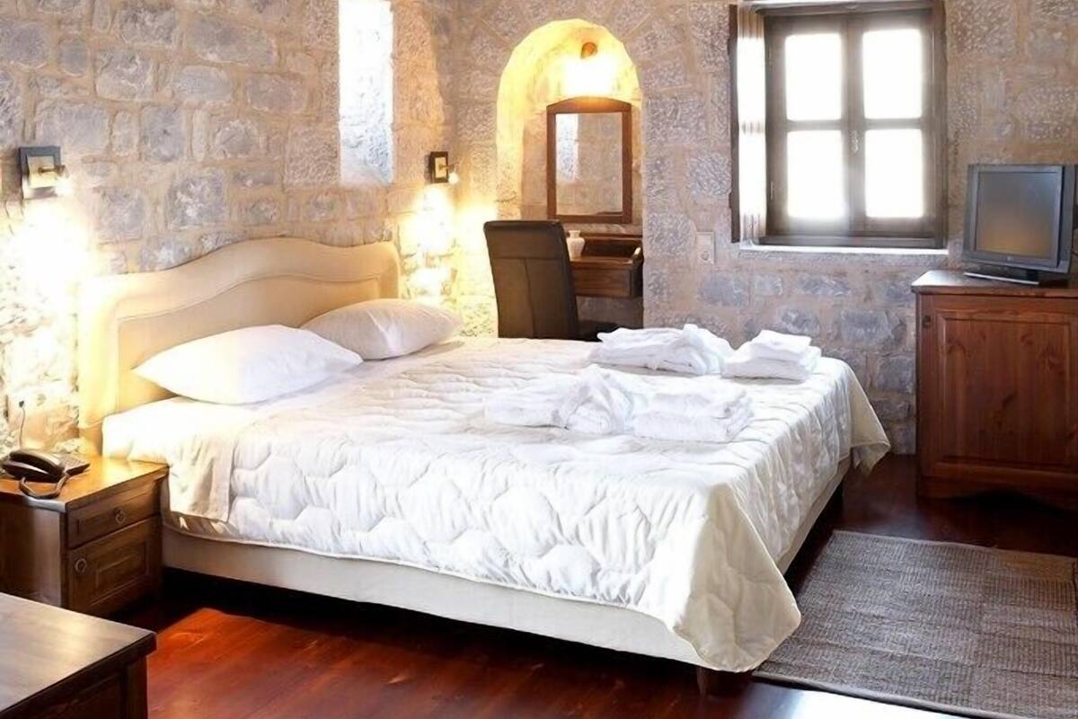 A bedroom with a stone wall, wooden ceiling beams, a bed with white bedding, a bedside table, a chair, a TV, and a window with natural light.