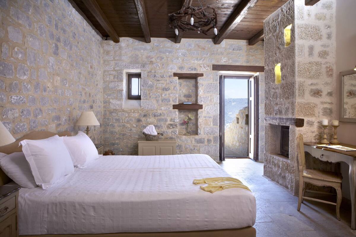 A bedroom with stone walls, a bed with white linens, a nightstand, a lamp, a small table with a basket, and a chair.