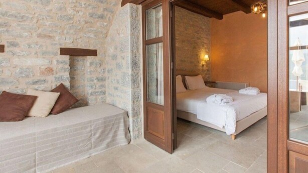 Une chambre avec un mur en pierre, une porte en bois, un lit avec du linge de lit blanc et une table de chevet avec une lampe.
