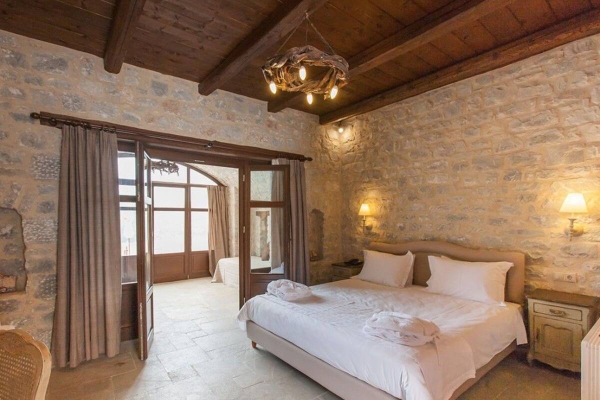 A bedroom with a stone wall, wooden ceiling beams, a bed with white linens, and a chandelier.
