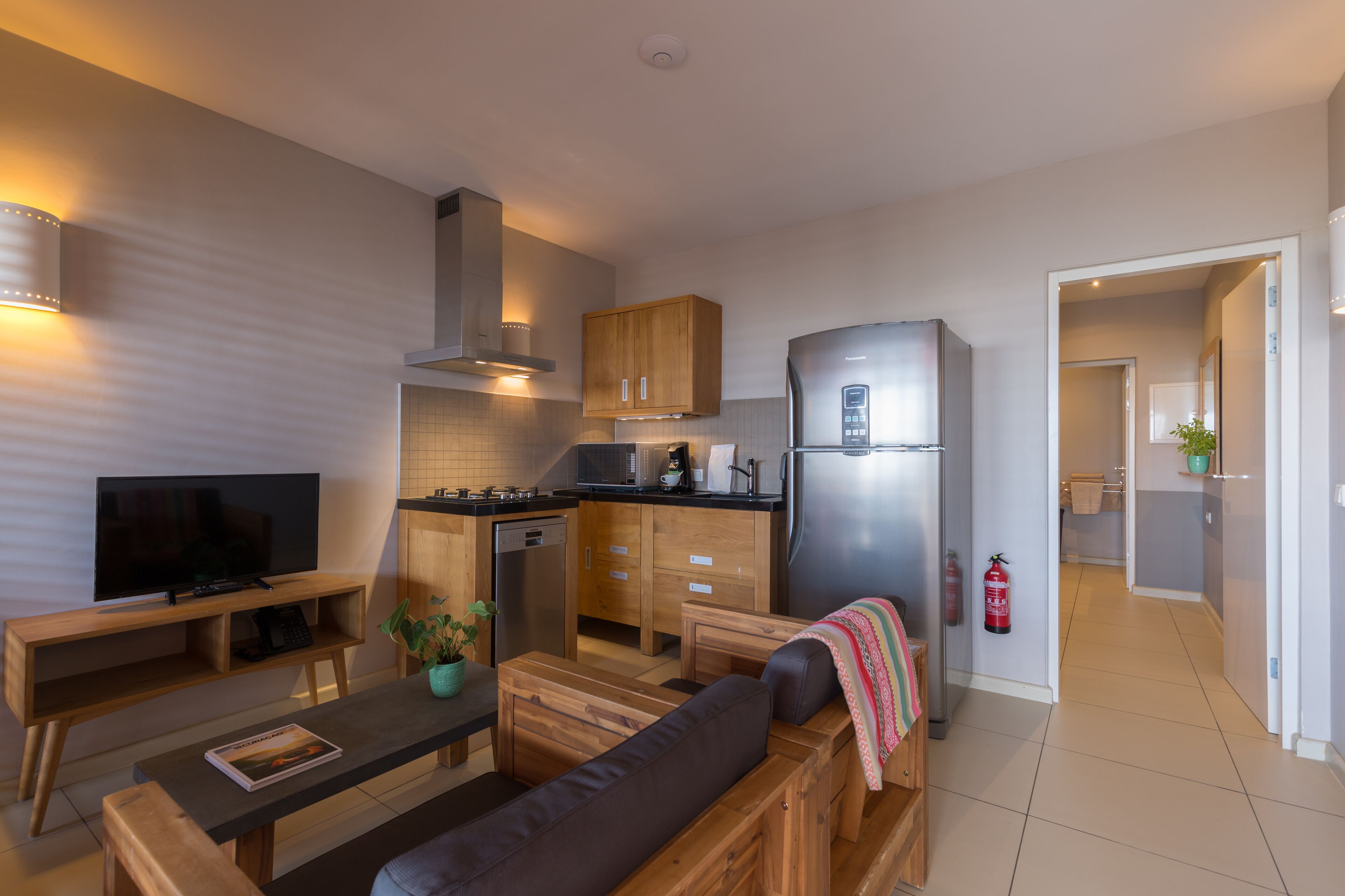 A modern kitchen with wooden cabinets, a stainless steel refrigerator, and a built-in oven. There is a dining area with a wooden table and a sofa.