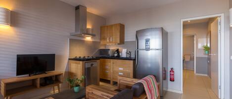 A modern kitchen with wooden cabinets, a stainless steel refrigerator, and a built-in oven. There is a dining area with a wooden table and a sofa.