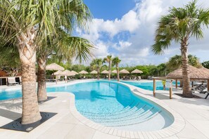 A swimming pool with palm trees and thatched umbrellas, surrounded by lounge chairs and a clear sky. - Morena Resort (Jan Thiel)