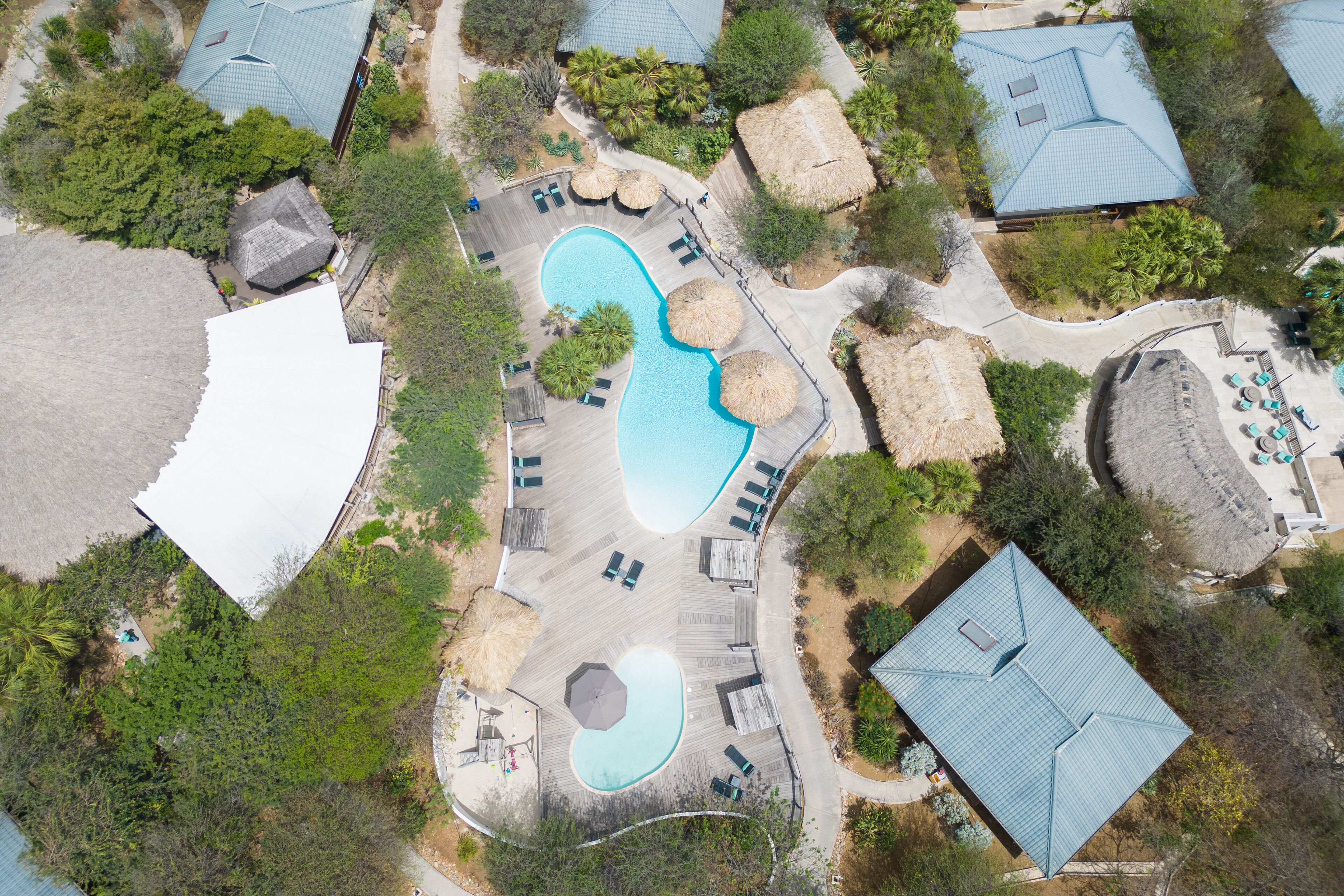 Eine Vogelperspektive zeigt einen luxuriösen Urlaubsort mit einem großen Schwimmbecken, Liegestühlen und Bungalows mit Strohdächern.