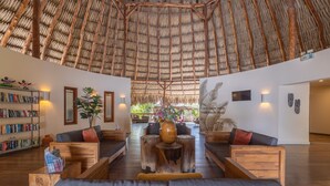 A spacious lobby with a high thatched ceiling, comfortable seating, a central wooden coffee table, and a bookshelf. - Morena Resort (Jan Thiel)