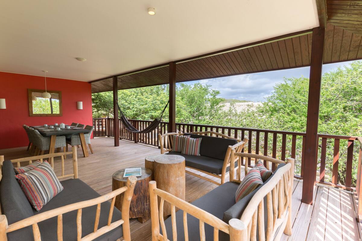 A covered outdoor seating area with wooden furniture, a dining table, and a hammock.