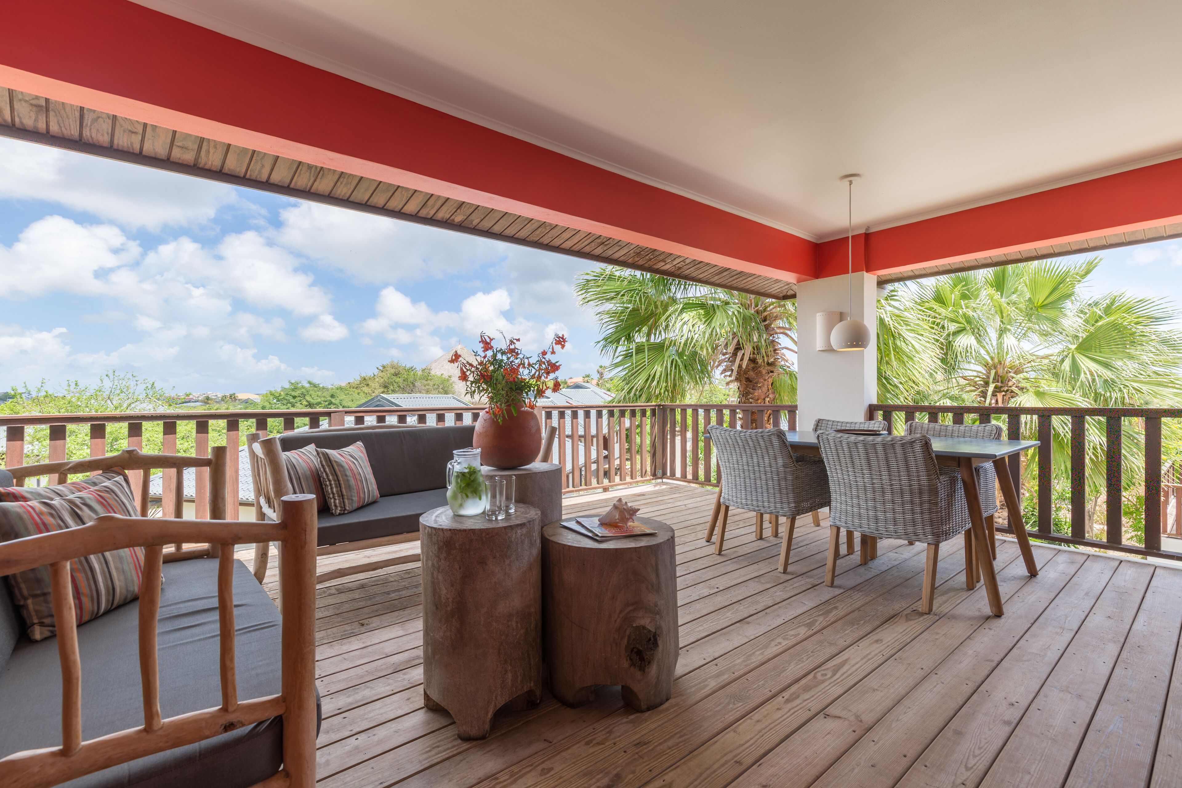 Eine Holzterrasse mit rotem Dach, Sitzgelegenheiten im Freien und Blick auf Palmen.