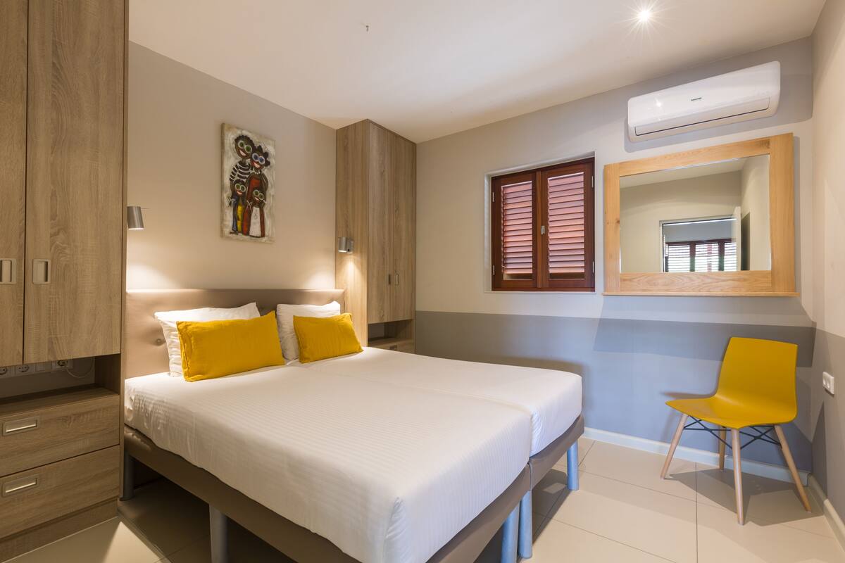 A bedroom with a bed, bedside lamps, a chair, a wall-mounted air conditioner, and a mirror.