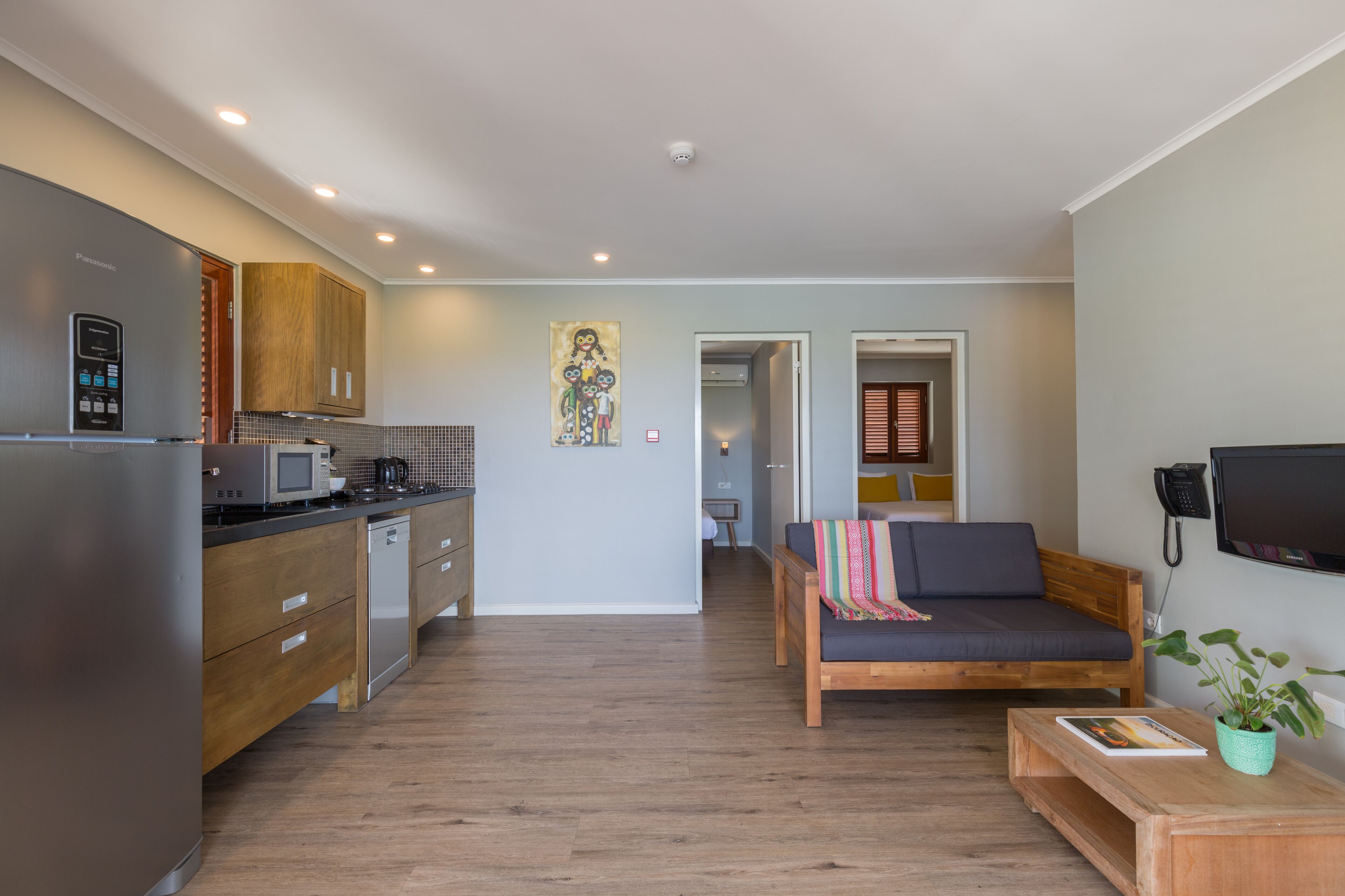 A modern kitchen with a built-in microwave, wooden cabinets, and a stainless steel refrigerator. There is a wooden bench with a cushion and a small table with a plant and a magazine.