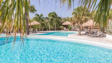A clear blue pool surrounded by palm trees and thatched umbrellas, with lounge chairs and a fence in the background.