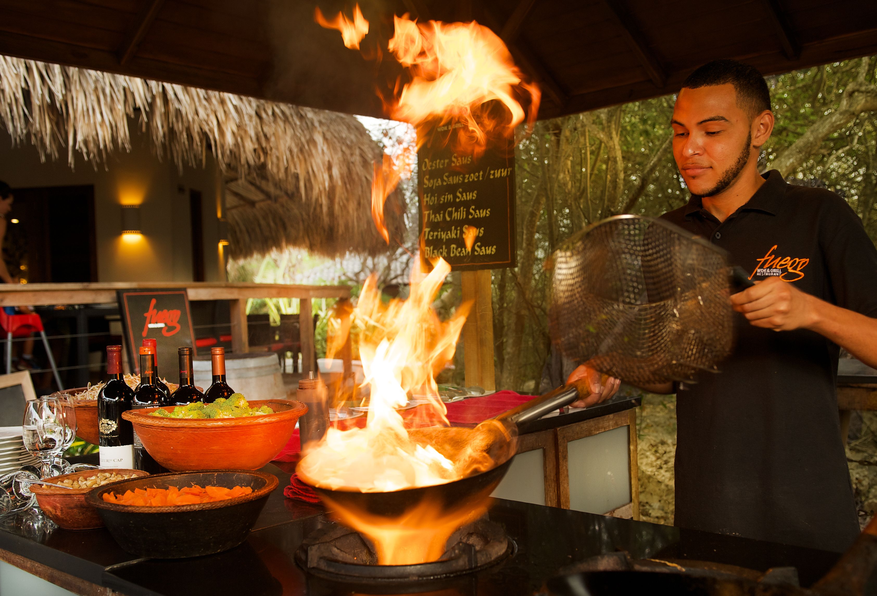 Un chef cocinando con llamas en un wok, rodeado de diversos ingredientes y utensilios de cocina.
