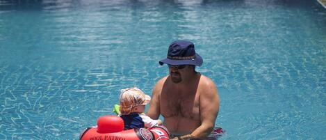 A man in a pool with a child on a float, surrounded by other people and a beach in the background.