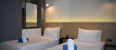 A hotel room with two beds, each with white linens and blue pillows, a wooden headboard, and a large mirror on the left wall.