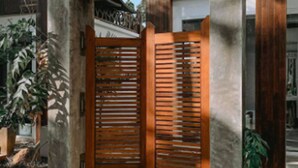 Une entrée en béton avec une porte en bois, entourée de plantes et un bâtiment moderne en arrière-plan.