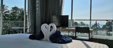 A hotel room with a large bed, a TV, a desk, and a view of palm trees and the ocean.