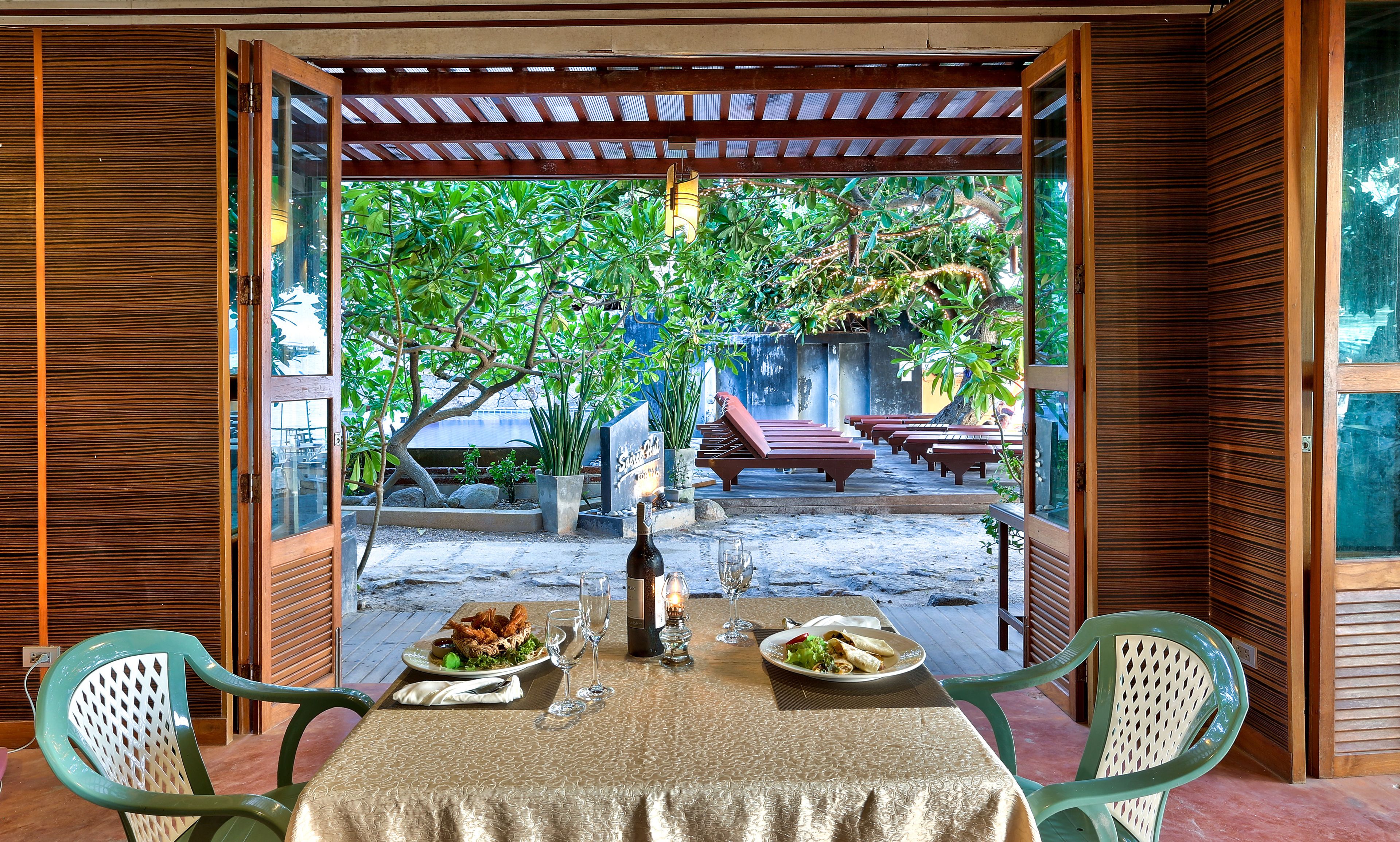 An outdoor dining area with a table set for two, a bottle of wine, and a candle. There are chairs and a bench in the background.