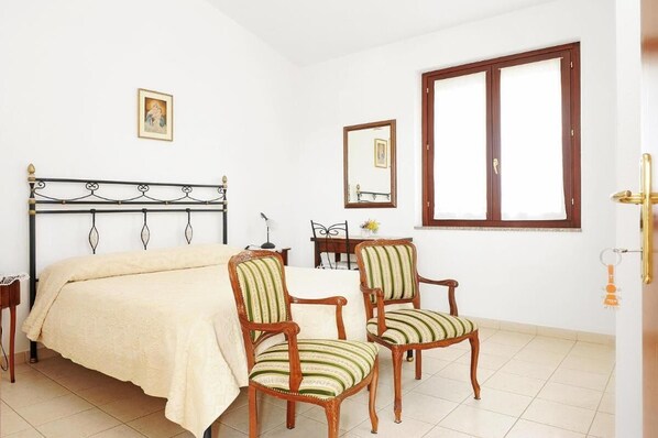 A bedroom with a bed, two upholstered chairs, a wooden nightstand, a desk with a lamp, a mirror, and a window with curtains. - Il Giardino di Valentina (Escolca)