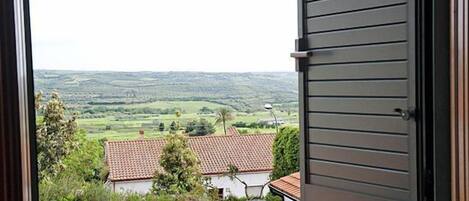 Uitzicht door een open raam op een landelijk landschap met glooiende heuvels, een wit huis met een rode dakbedekking en een metalen hek.