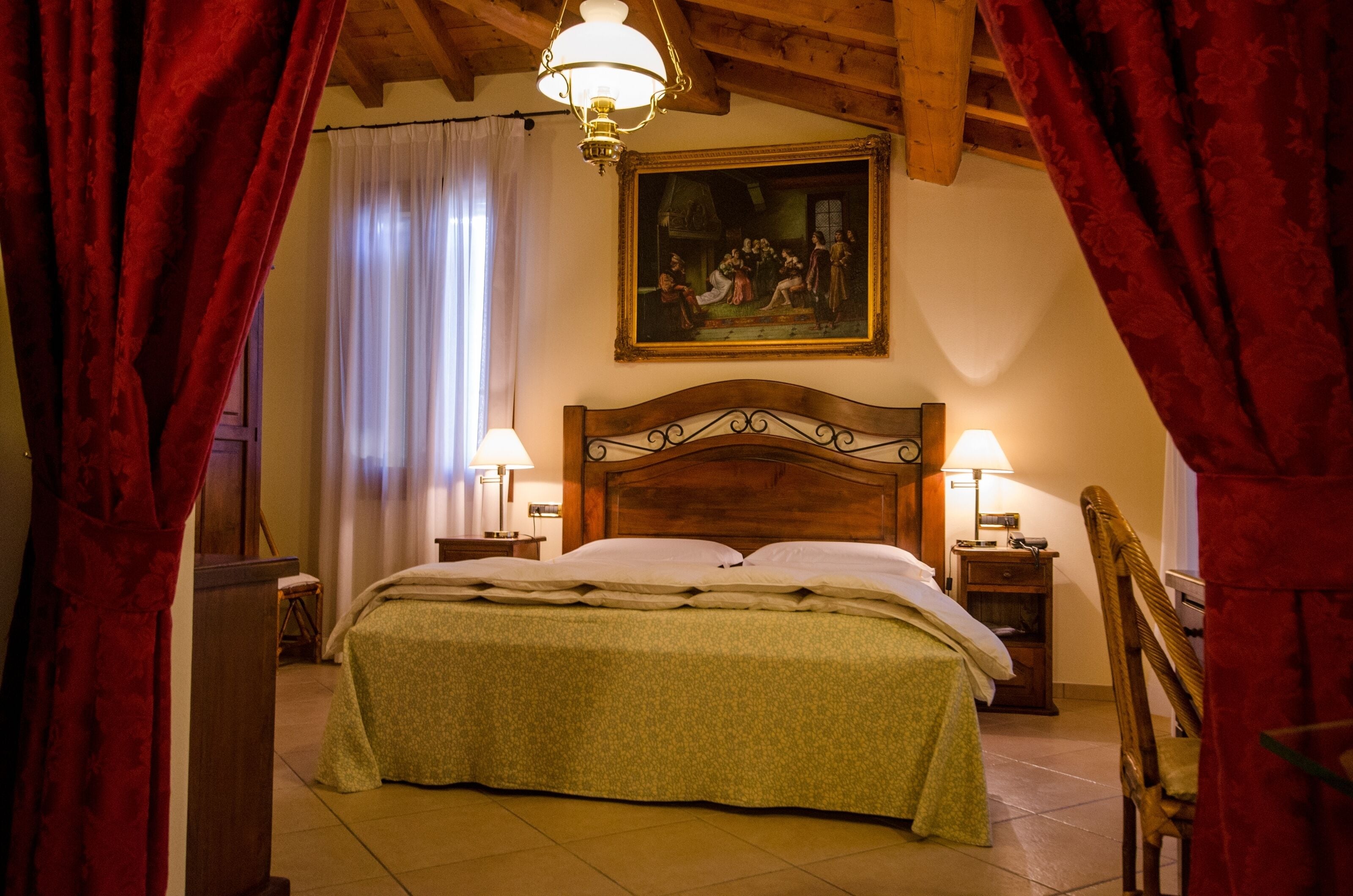 A bedroom with a wooden headboard, two bedside lamps, a bed with a green bedspread, a painting above the bed, and red curtains framing the window.
