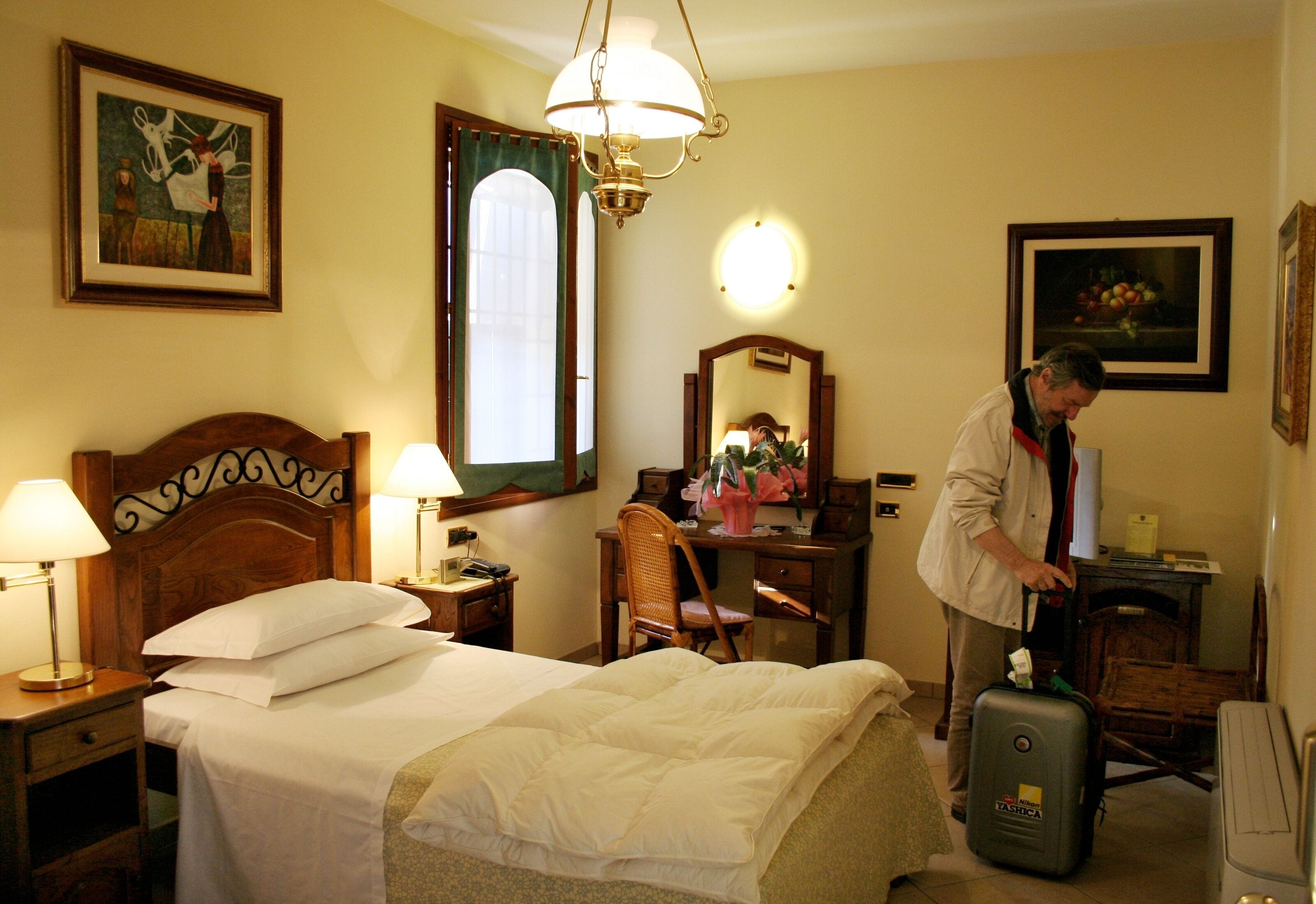 A hotel room with a bed, a desk, a chair, a suitcase, and a mirror.