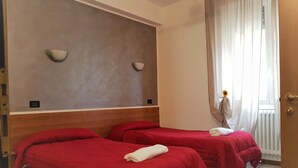 Une chambre d’hôtel avec deux lits, chacun recouvert d’une couverture rouge et agrémenté d’une serviette blanche. On y trouve une tête de lit en bois, un luminaire fixé au mur et une fenêtre avec des rideaux.